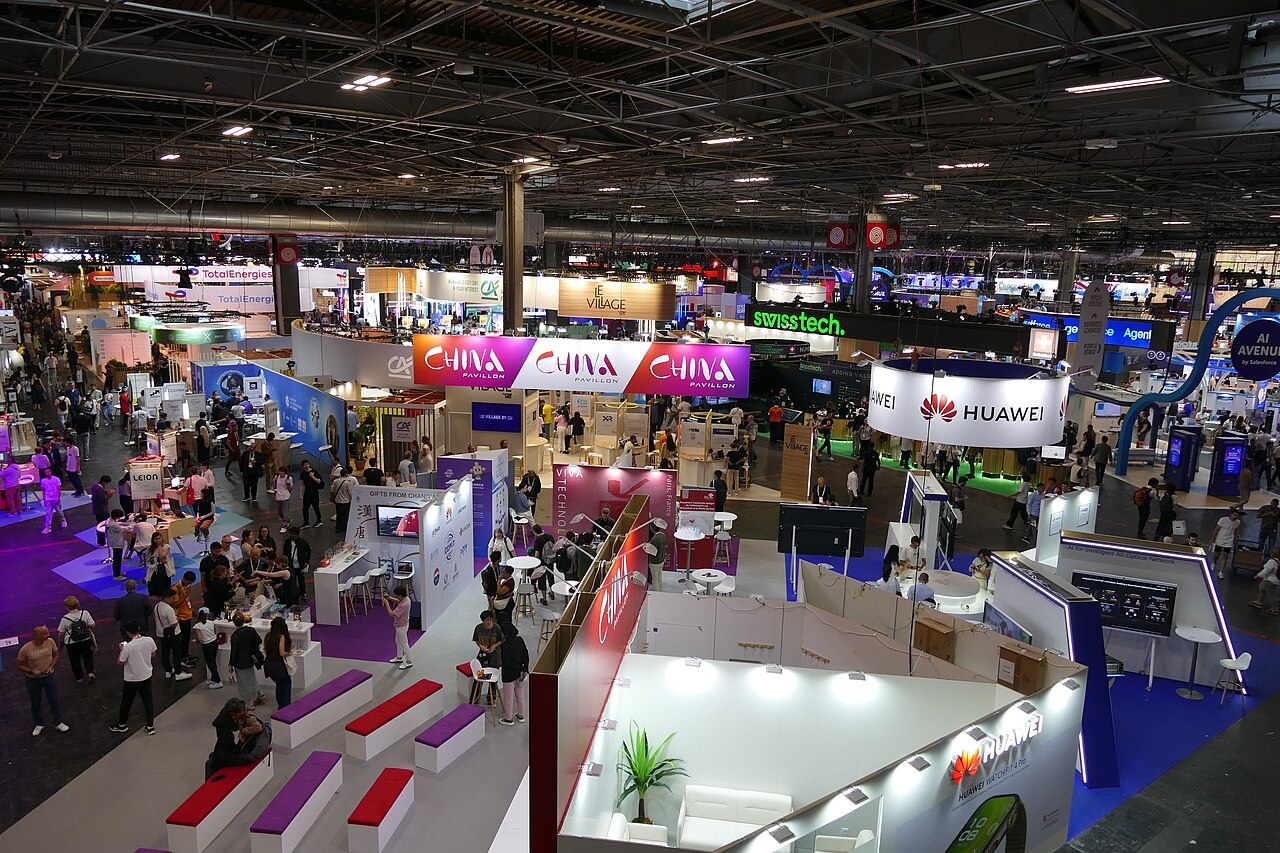 Vue plongeante sur un hall animé de Paris Expo Porte de Versailles pendant le salon VivaTech : allées parcourues de visiteurs entre les stands lumineux des exposants, avec les bannières de pavillons collectifs (China Pavillon, Le Village, swisstech, Huawei, AI Avenue) suspendues au-dessus des allées